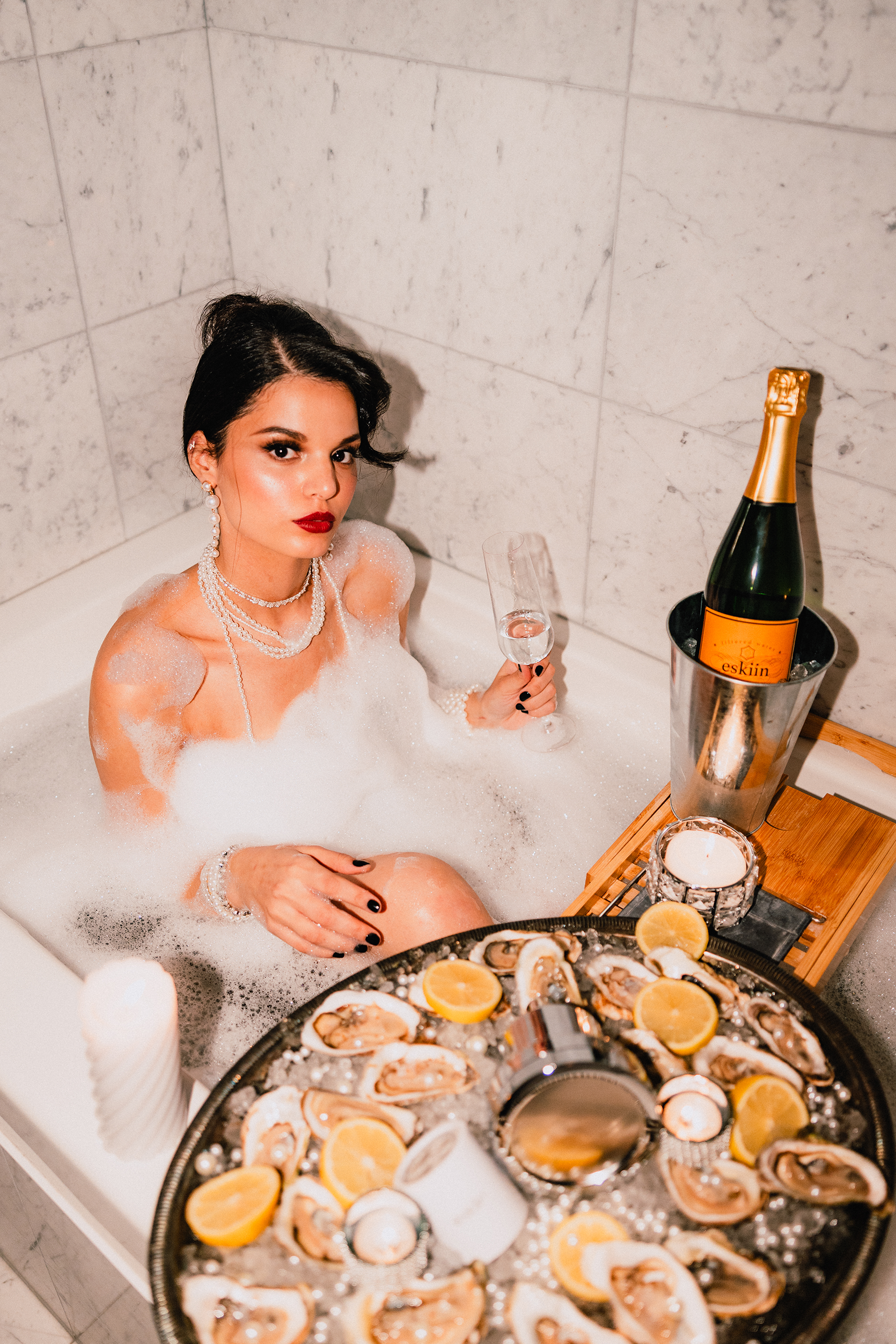 Woman in bathtub with champagne, pearls, and seafood platter.